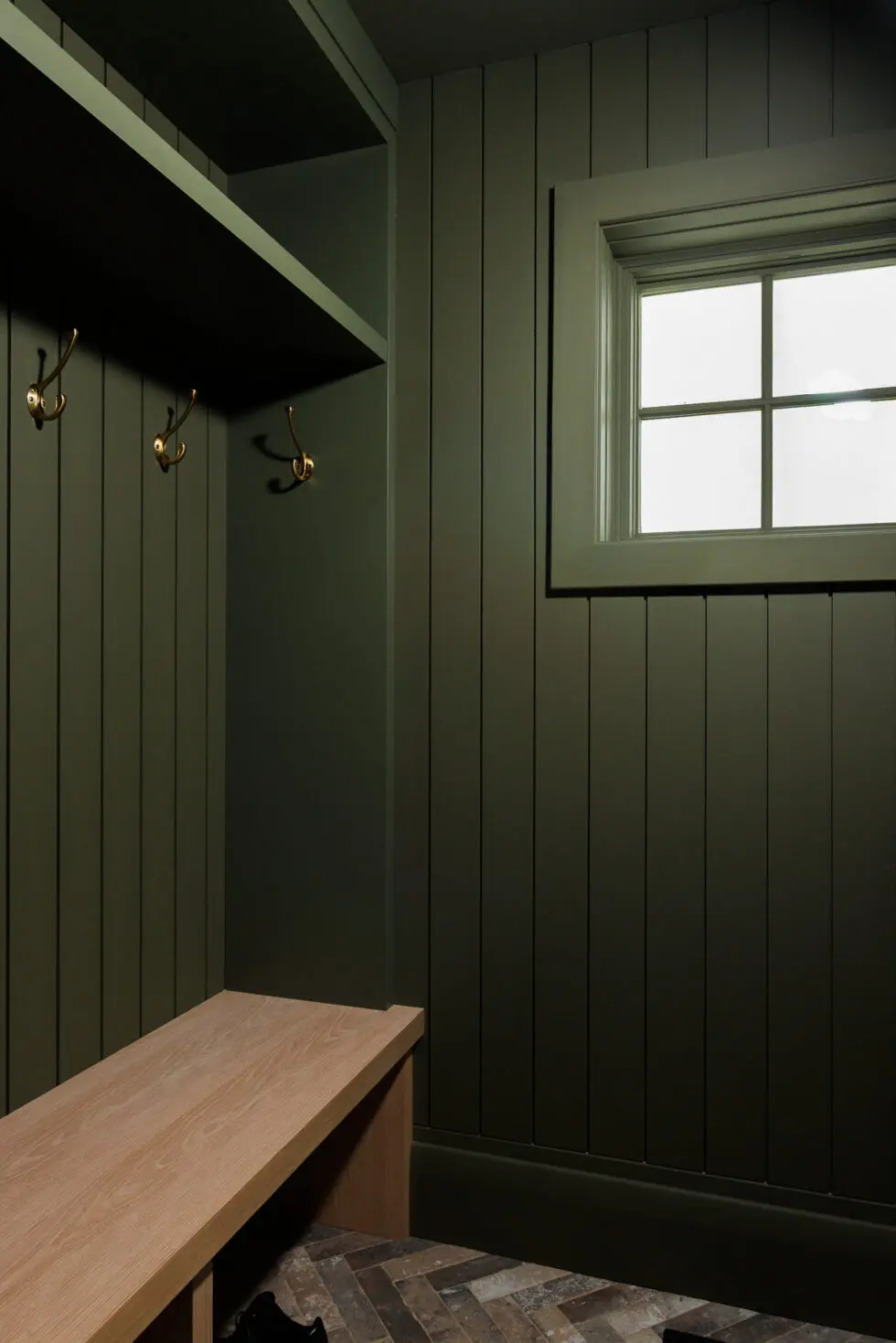 Stylish mudroom or entryway with dark olive green vertical shiplap walls, a light wood built-in bench, and brass coat hooks.