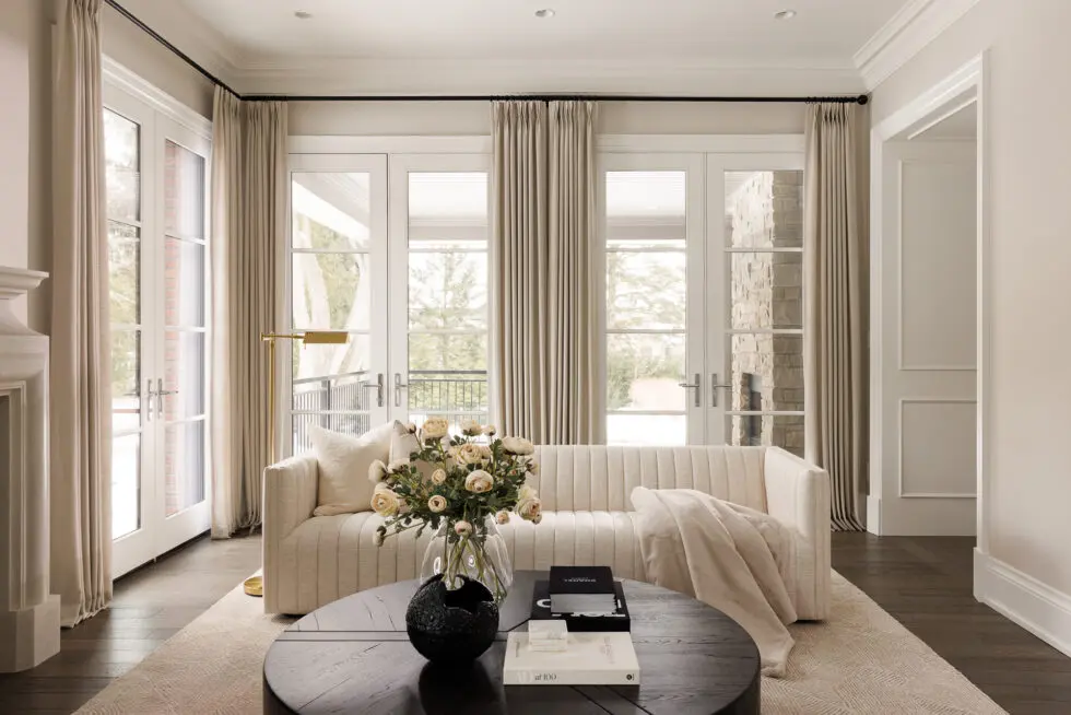Modern neutral living room with cream channeled sofa, black coffee table, large French doors, and beige drapes.