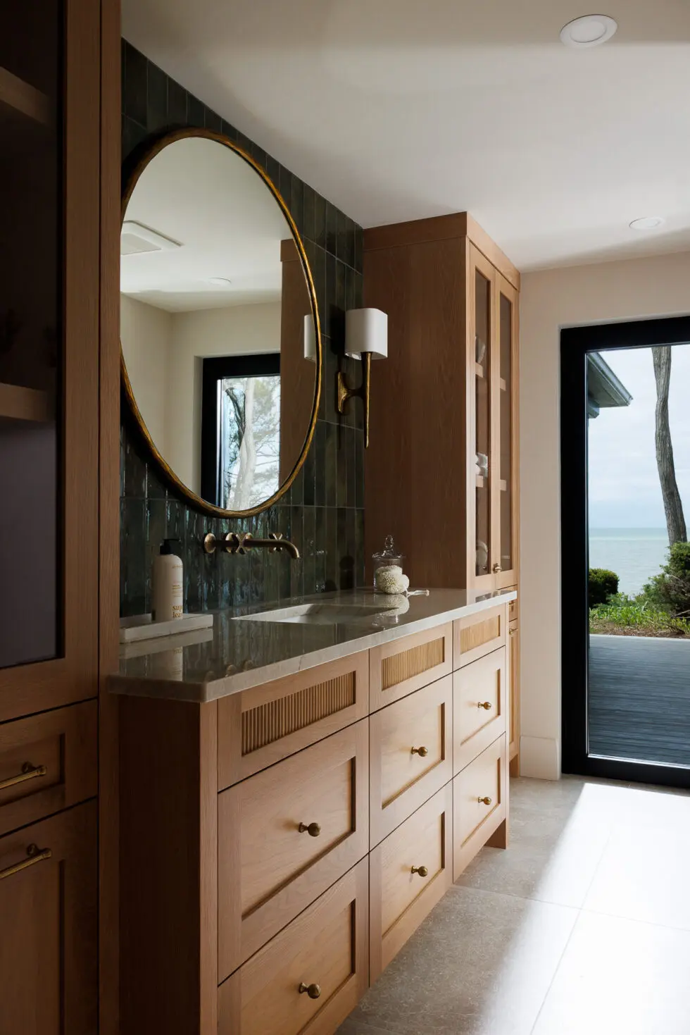 Modern bathroom with a light wood vanity, round gold-framed mirror, dark green subway tile accent wall, brass wall-mounted faucet, and a large glass door opening to an ocean view.