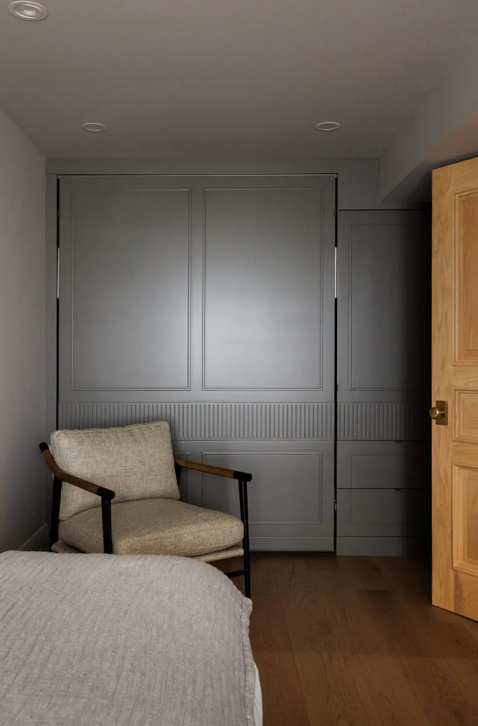 A modern bedroom features a large gray paneled built-in wardrobe with decorative reeding, a beige upholstered armchair, and warm hardwood flooring.