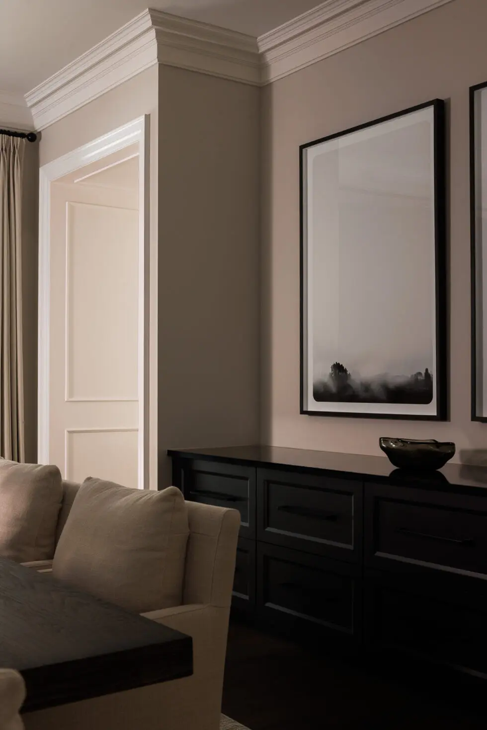 Elegant interior featuring a black console table, minimalist black and white landscape art, beige upholstered seating, and classic white crown molding.