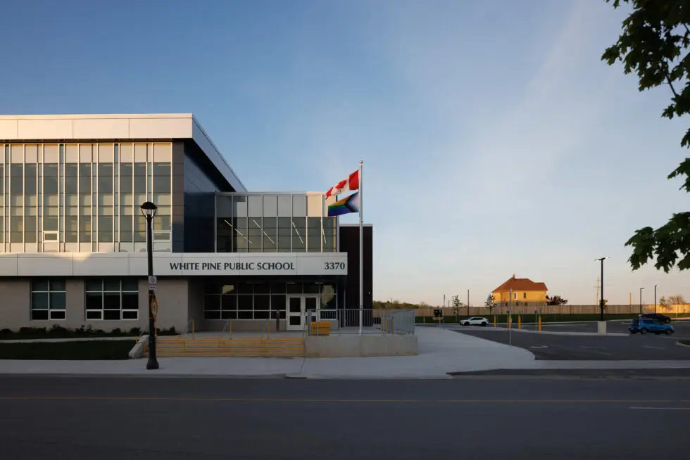 White Pine Public School building with Canadian and Progress Pride flags flying, modern architecture, and a street in front under a clear sky.