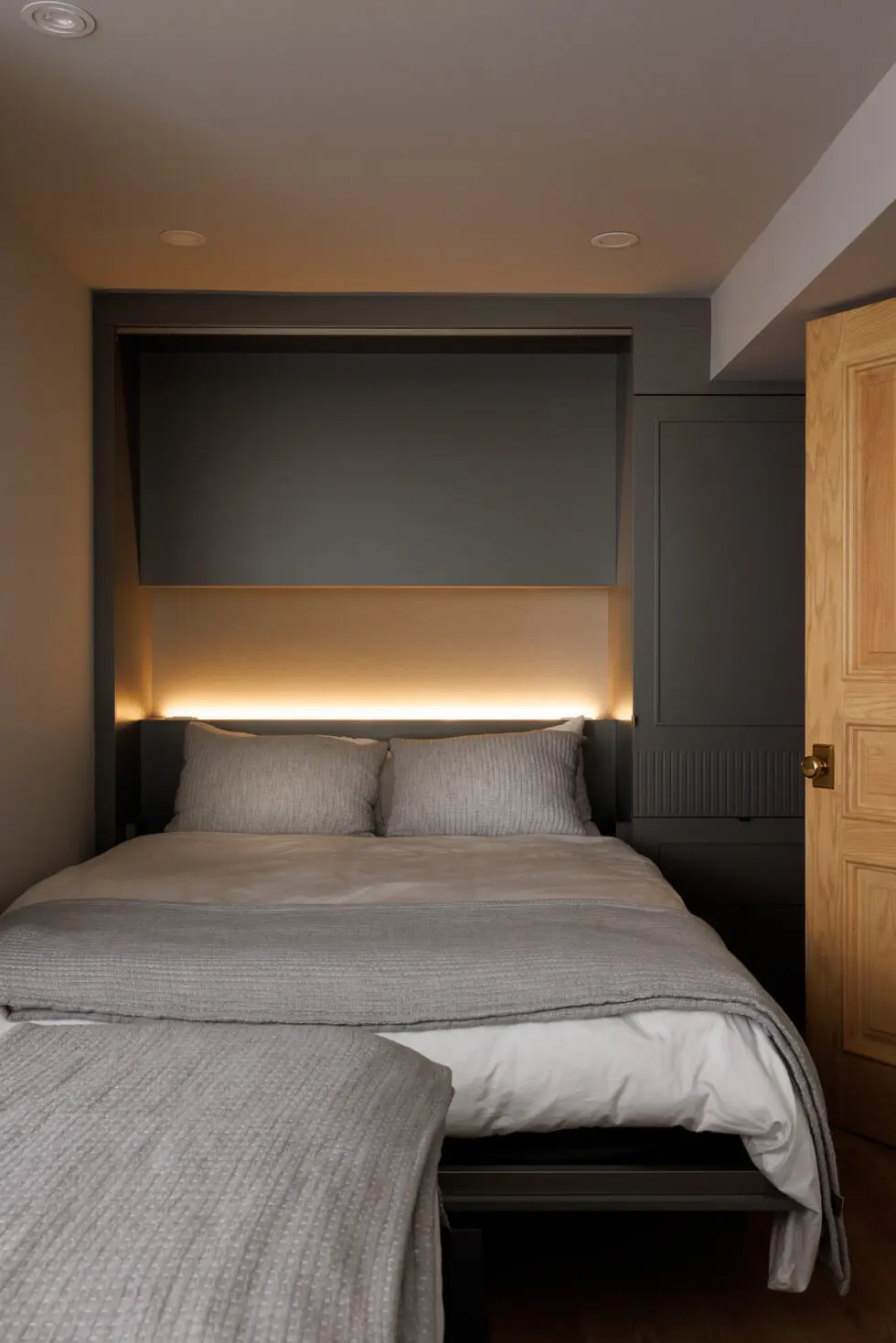 Modern compact bedroom with grey bedding, warm LED headboard lighting, and built-in dark grey storage.