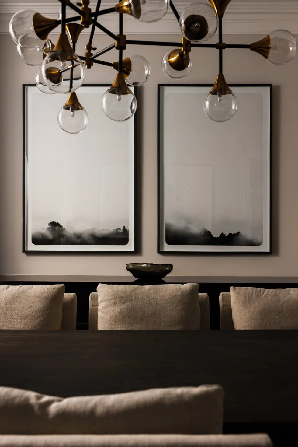 Modern brass and glass chandelier illuminating a stylish dining room with two framed misty landscape artworks, a dark wood table, and beige upholstered chairs.