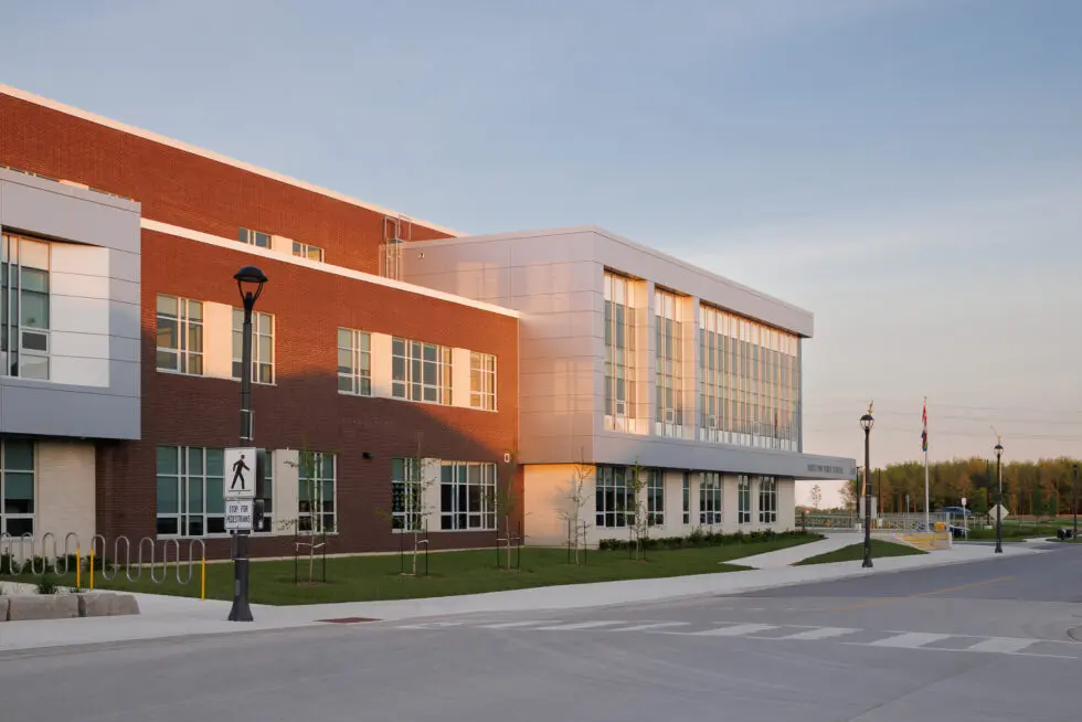Exterior view of White Pine Public School, a modern building with red brick and metallic grey facade, under a clear sky during golden hour. The foreground features a sidewalk, street, bike racks, and a pedestrian crossing sign.