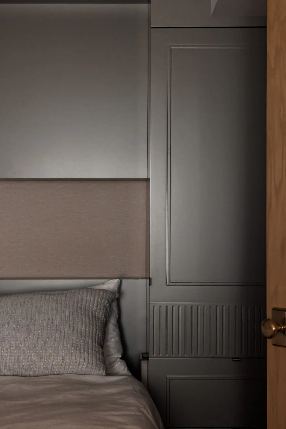 Modern bedroom interior with grey bedding, a sophisticated upholstered headboard, and dark grey built-in cabinetry next to a wooden door with a brass doorknob.