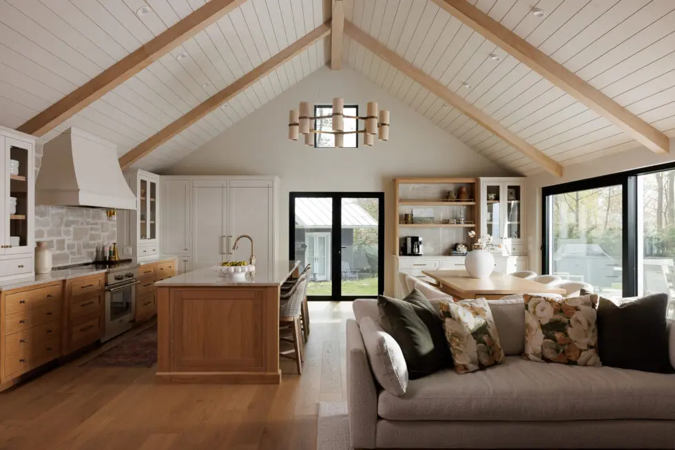 Spacious open concept kitchen and living room with vaulted ceilings and wood beams, featuring a large kitchen island, light wood cabinetry, stone backsplash, and a modern living area with a sofa and floral pillows. Large glass doors open to the outdoors.