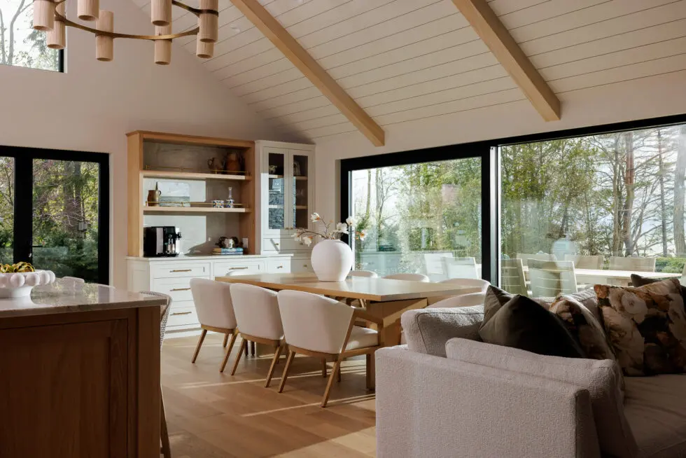 Modern open concept living and dining room with vaulted ceiling, light wood beams, a contemporary chandelier, and a built-in buffet. The space features a wood dining table with armchairs, a plush sectional sofa, and expansive windows overlooking a natural landscape.