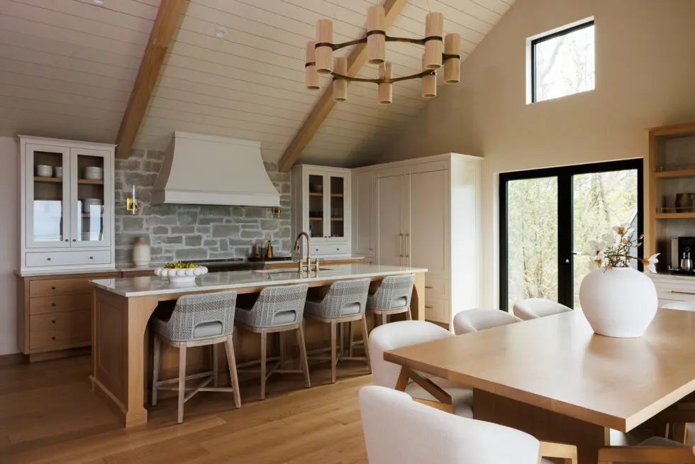 Modern open concept kitchen and dining area featuring a large wooden island with bar stools, stone accent wall, white cabinetry, a vaulted wood plank ceiling with chandelier, and a natural wood dining table with upholstered chairs.