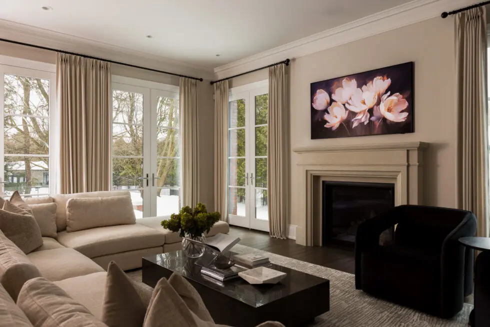 A modern and luxurious living room featuring a large beige sectional sofa, a stone fireplace with a floral art piece, a dark coffee table with decor, and large windows with neutral drapes looking out to a winter landscape.