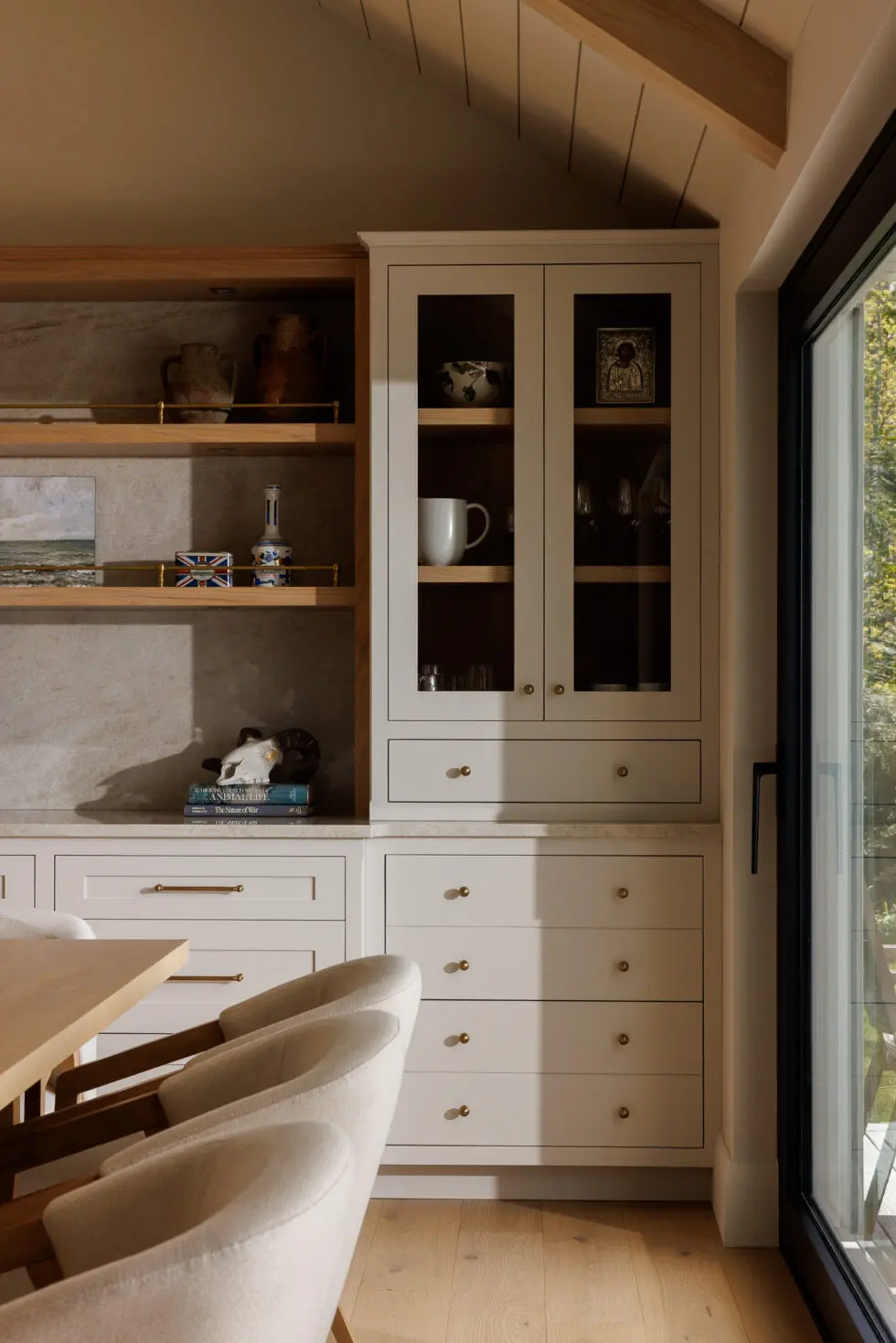 Modern dining room with custom light grey built-in cabinetry, open wood shelving, brass hardware, and upholstered dining chairs next to a large window.