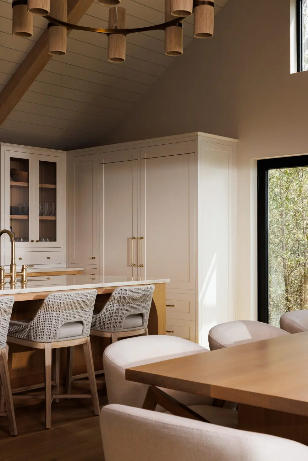 Modern open-concept kitchen and dining area with vaulted ceiling, light neutral cabinetry, kitchen island with woven bar stools, and a wooden dining table next to a large window.