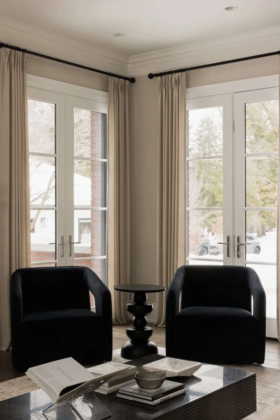 Elegant modern living room featuring two black velvet armchairs, a unique black sculptural side table, and a dark coffee table with decorative books, set against large French doors with natural drapes revealing a snowy outdoor scene.
