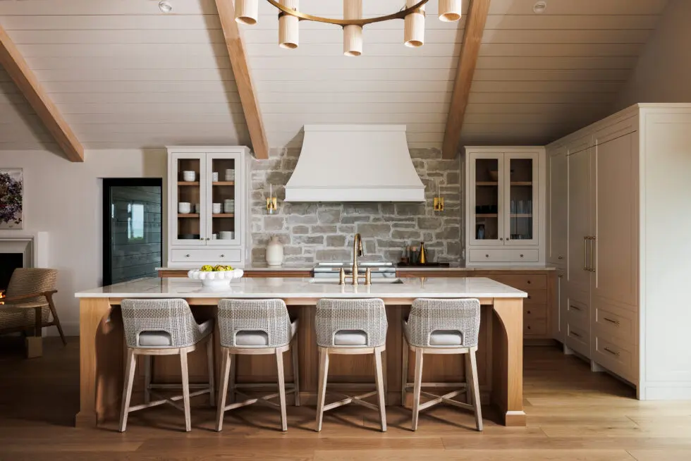 Modern farmhouse kitchen with a large wooden island, white quartz countertop, four woven bar stools, a stone backsplash, white custom range hood, and white Shaker-style cabinets. The kitchen features a vaulted ceiling with exposed wood beams and a contemporary brass chandelier.