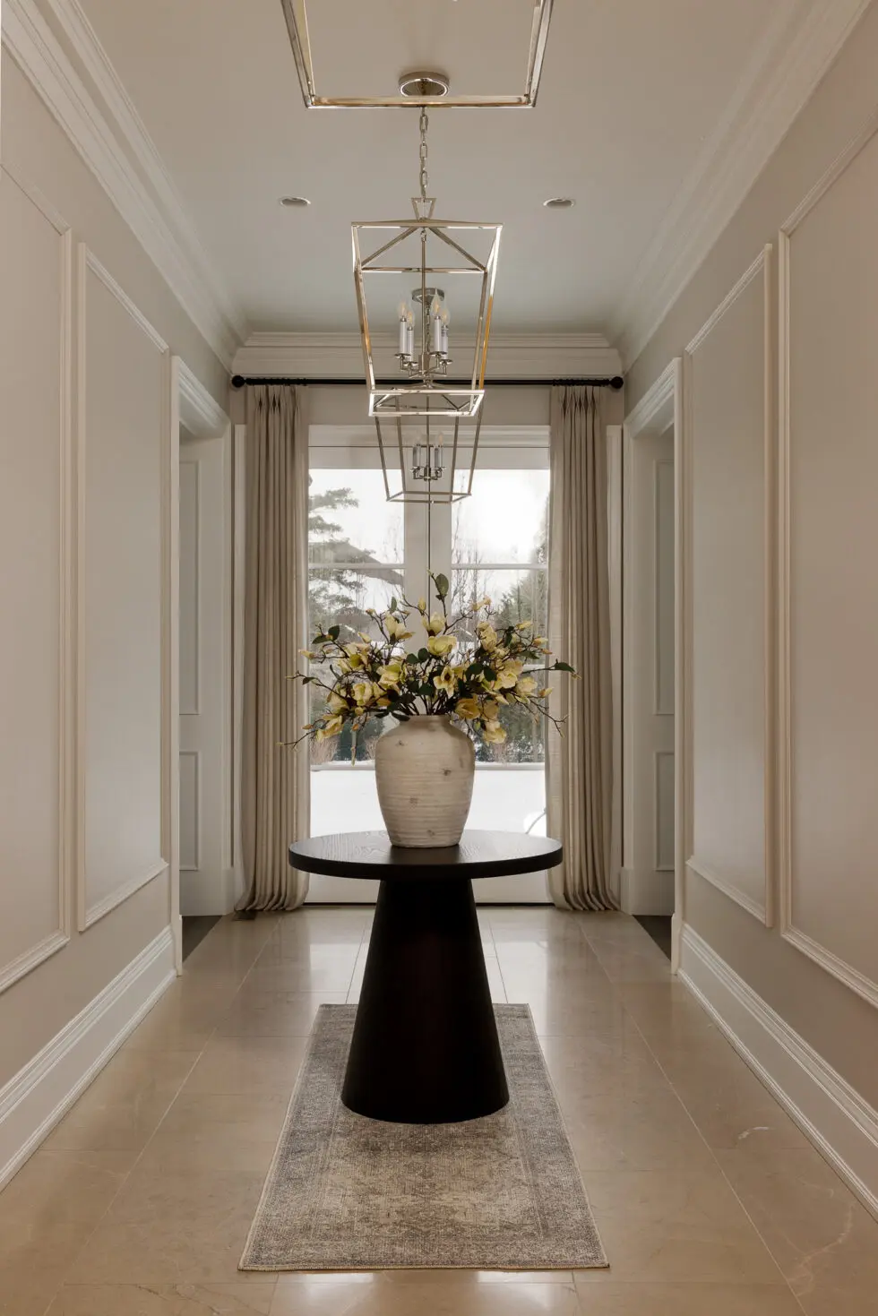 Luxurious hallway with dark round console table, large white vase with yellow artificial flowers, and elegant gold geometric chandeliers.