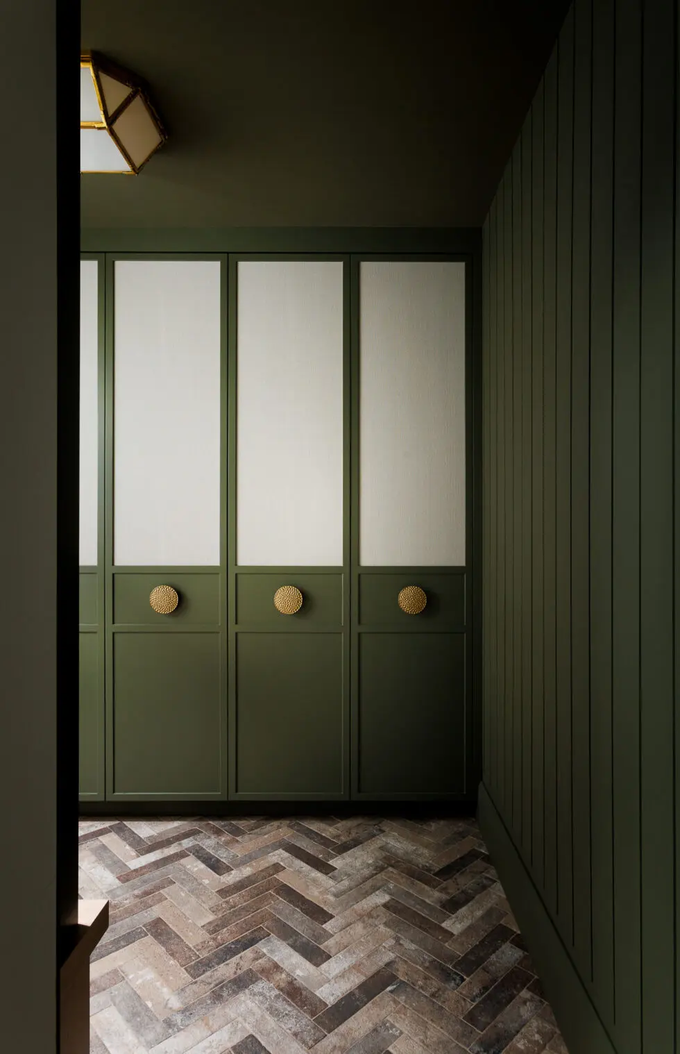 Elegant dark green built-in wardrobes with textured off-white inserts and gold brass handles, alongside a vertically paneled green wall and rustic herringbone tile flooring.