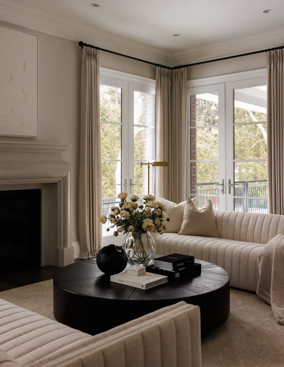 Modern luxury living room with a cream ribbed sofa, dark round coffee table, French doors, and a stone fireplace.