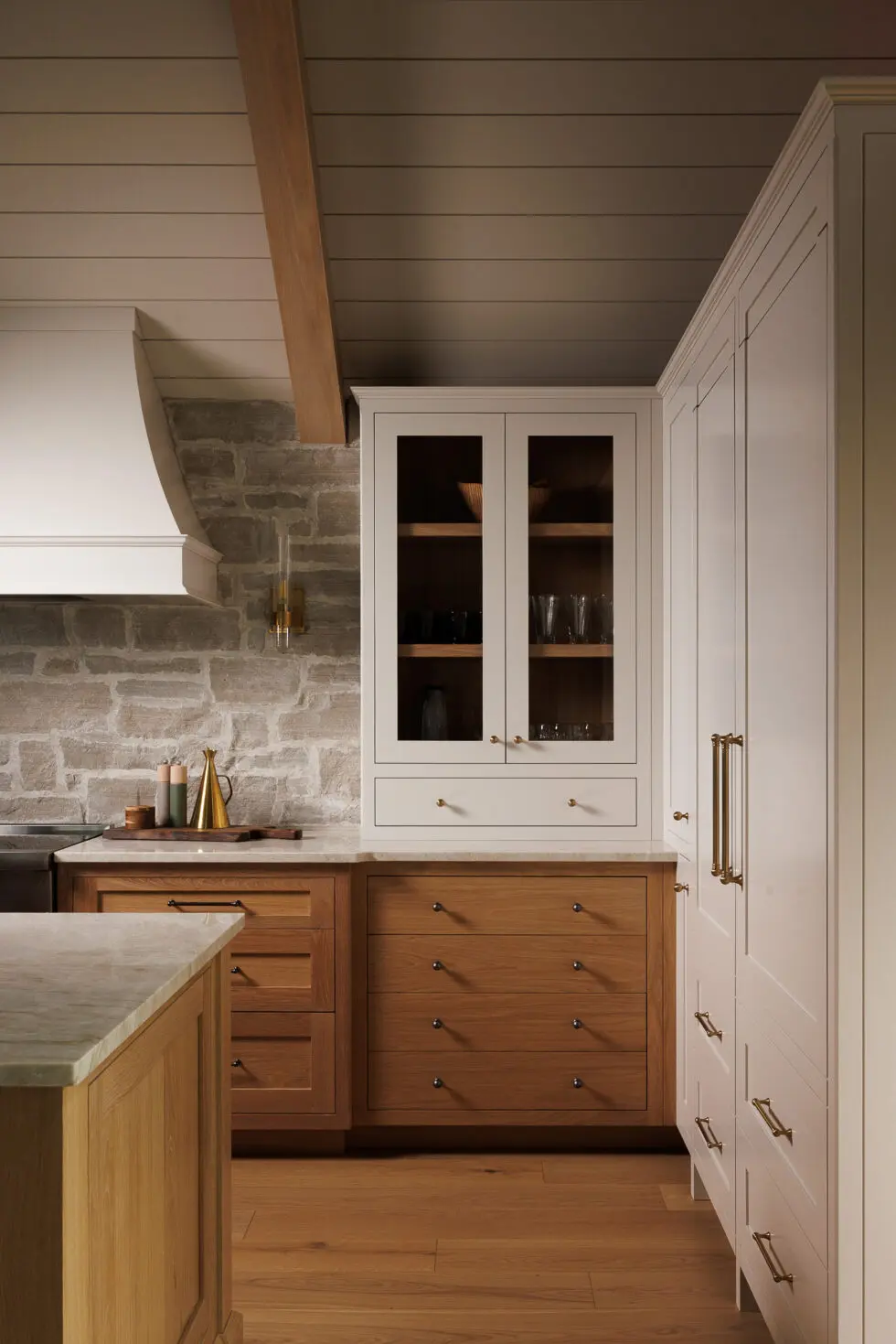 Modern transitional kitchen featuring cream upper cabinets, natural wood lower cabinetry and island, a stone backsplash, white range hood, wood beam ceiling, light wood flooring, and brass hardware with decorative accents.