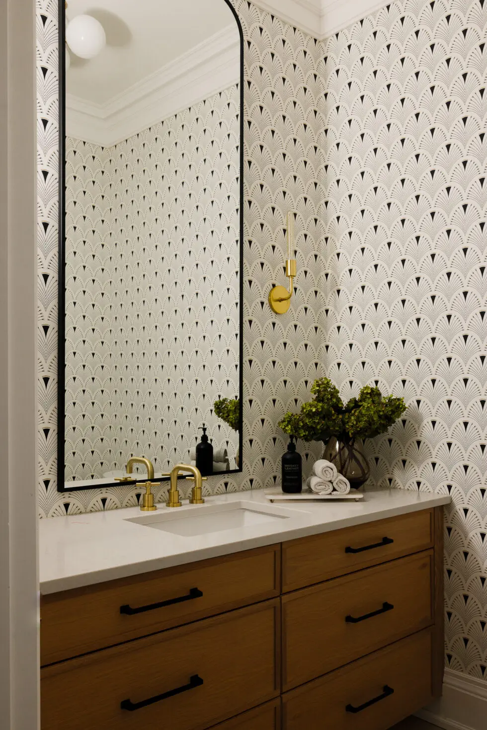 Modern bathroom with black and white Art Deco fan pattern wallpaper, arched mirror, wood vanity, and gold fixtures.