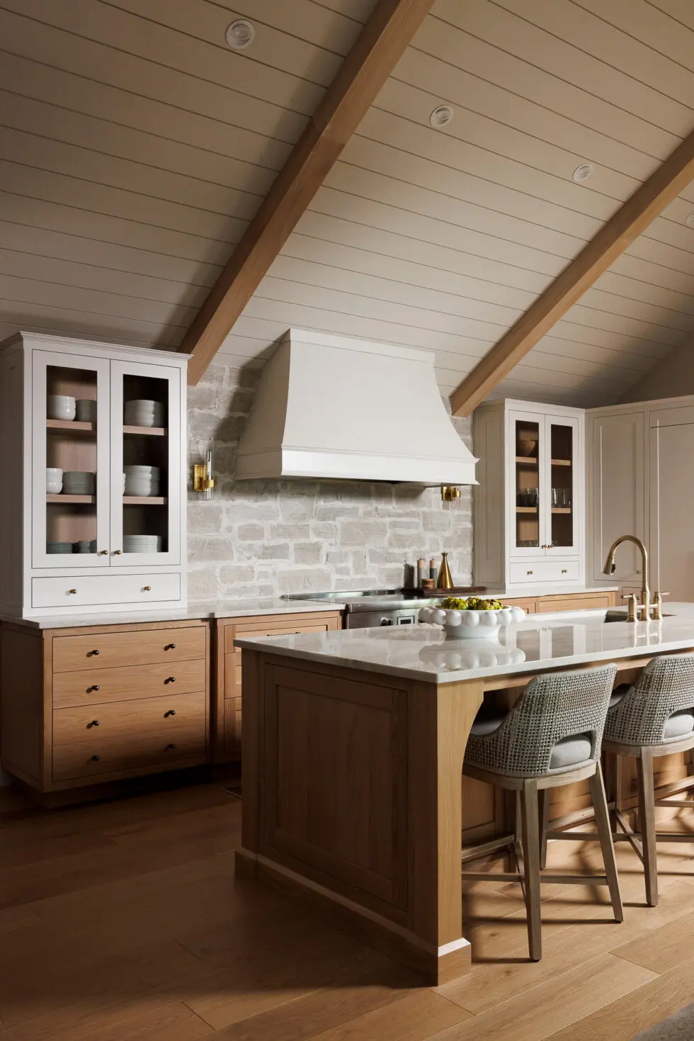 A spacious modern farmhouse kitchen featuring a large wooden island with white marble countertop and two woven bar stools, white upper cabinets, natural wood base cabinets, a stone backsplash, and a vaulted shiplap ceiling with exposed wood beams.