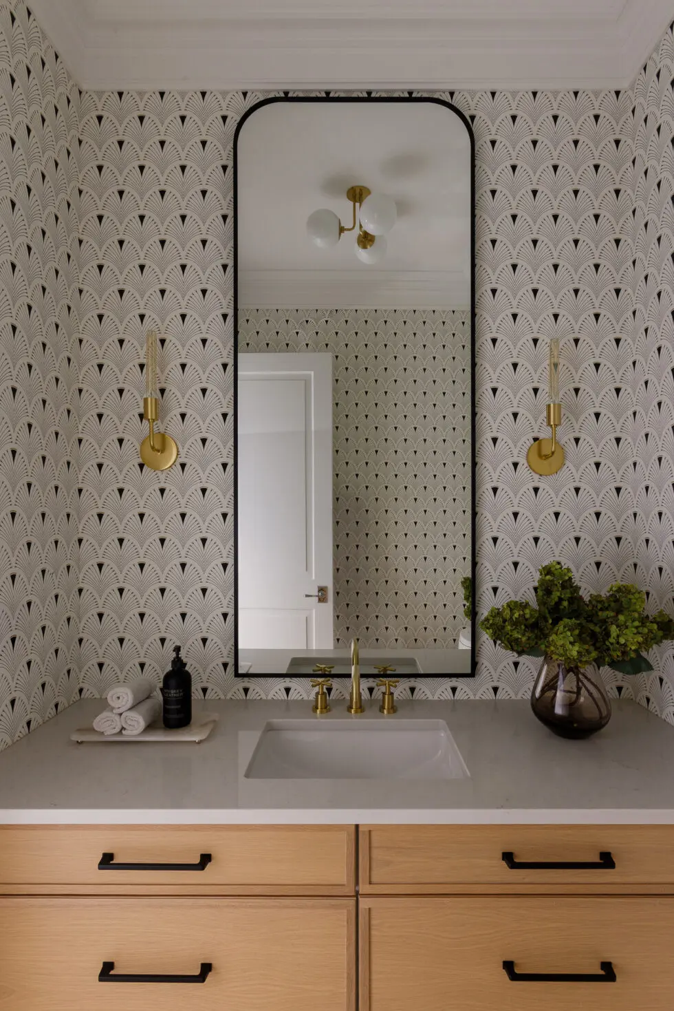Elegant powder room featuring black and white Art Deco fan wallpaper, a rectangular black-framed mirror, gold sconces, a light wood vanity with black hardware, and a white quartz countertop with a vessel sink and brass faucet.