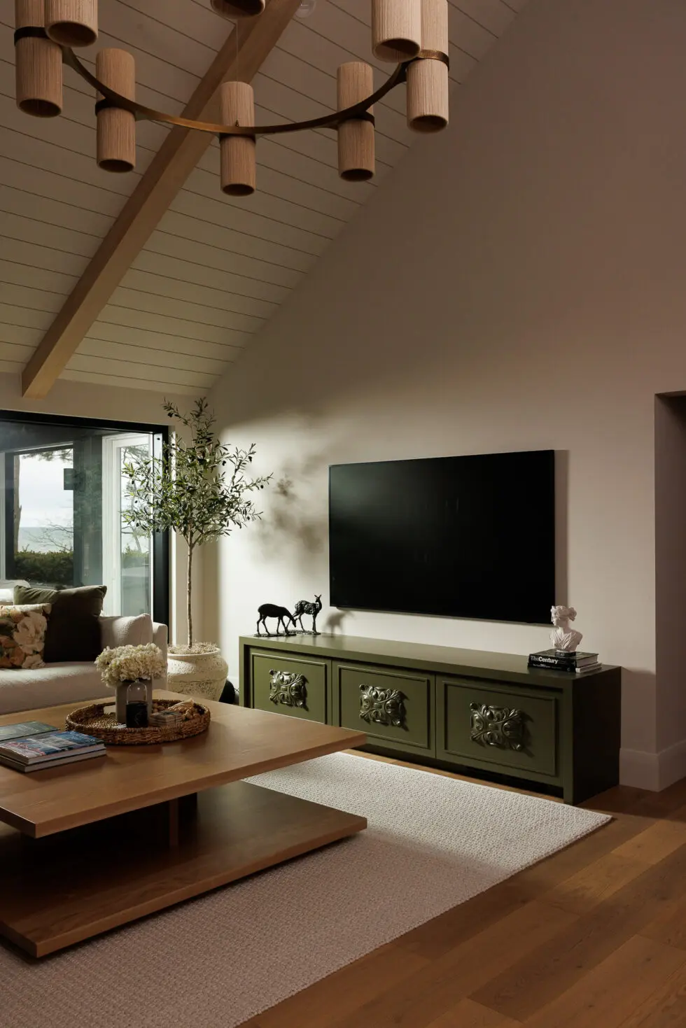 Modern living room with olive green media console, wood coffee table, and a large window overlooking a lake. The room features a vaulted ceiling, a unique chandelier, and a faux olive tree.
