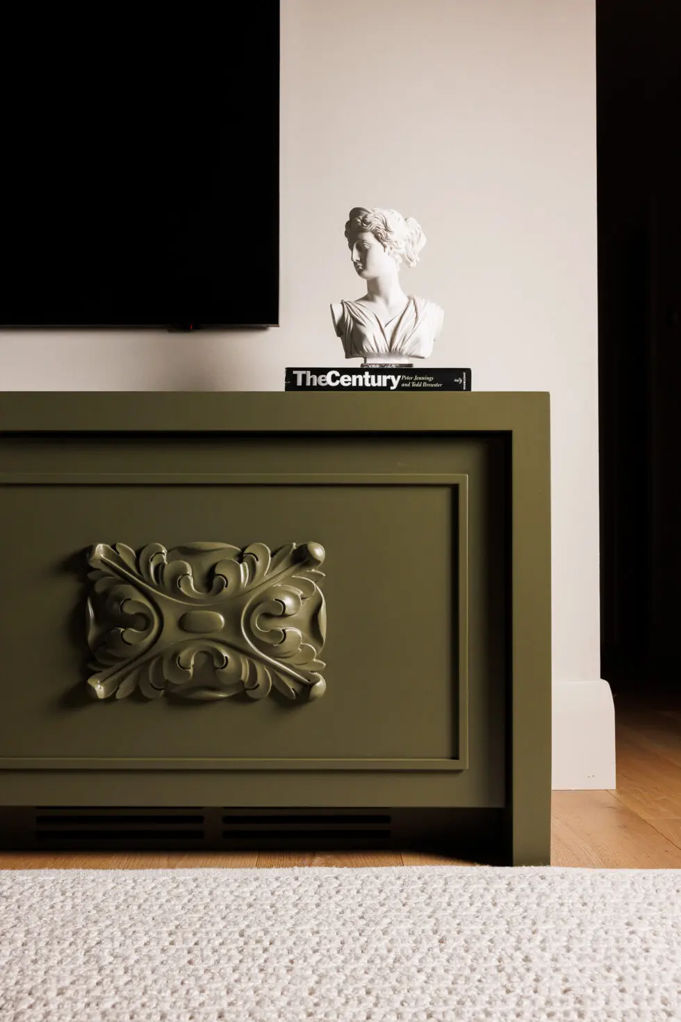 Olive green console cabinet with decorative molding, classical bust, and 'The Century' book under a mounted TV.