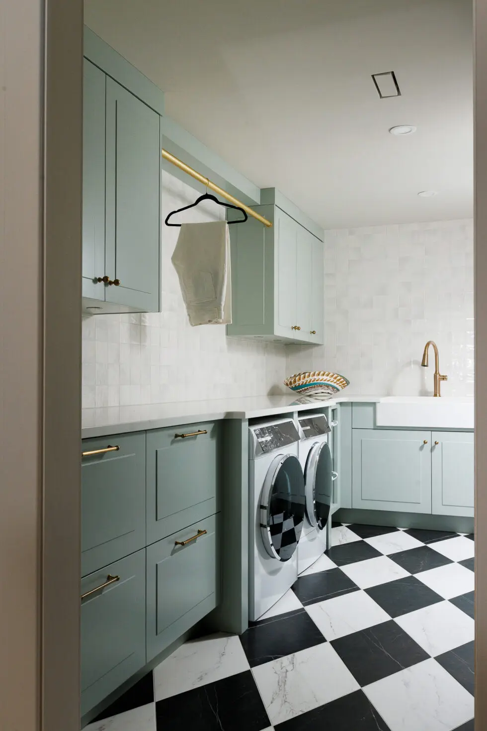 Modern laundry room with sage green shaker cabinets, brass hardware, black and white checkered floor, and front-load washer and dryer.