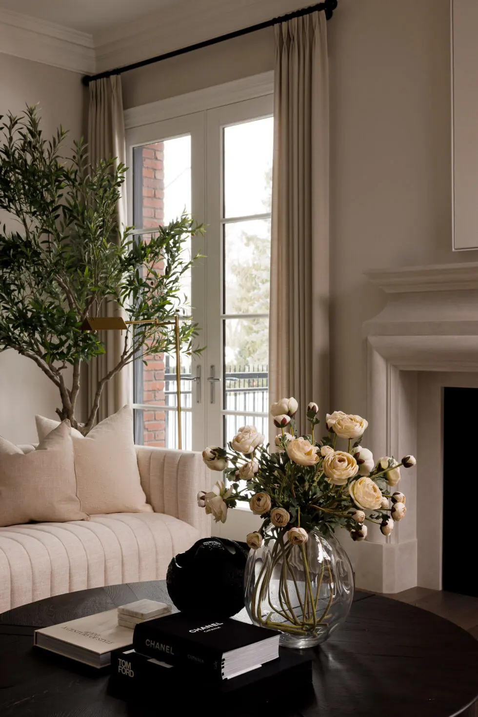 Elegant living room with a beige channel-tufted sofa, large faux olive tree, black coffee table, and a vase of cream ranunculus flowers by French doors.