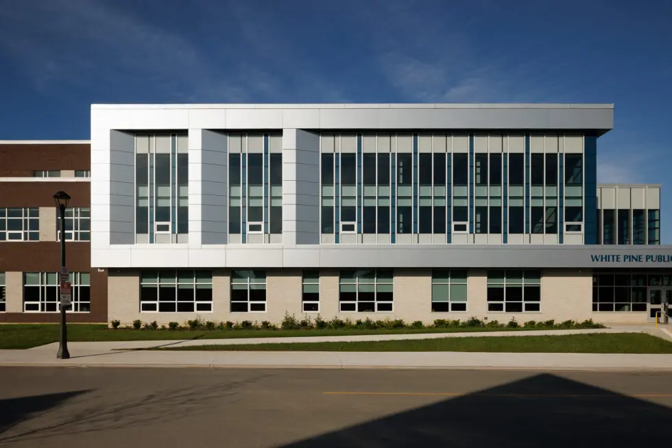 Front view of the modern White Pine Public School building under a clear blue sky, featuring a blend of brick and contemporary silver paneling with large windows, a sidewalk, and green grass.