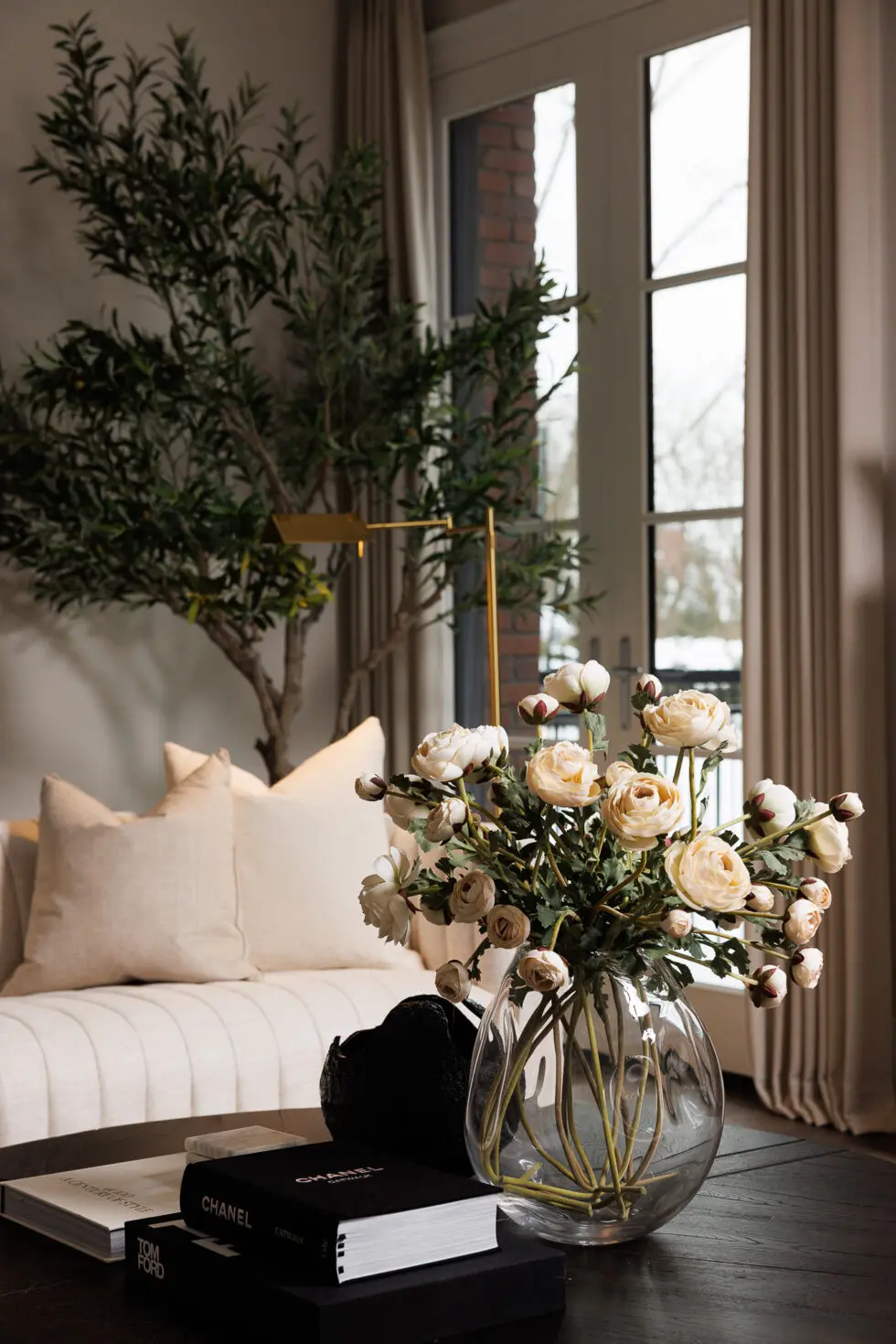 Elegant living room with a clear glass vase of artificial light pink ranunculus flowers on a dark wood coffee table, alongside fashion books, a cream tufted sofa, and an artificial olive tree.