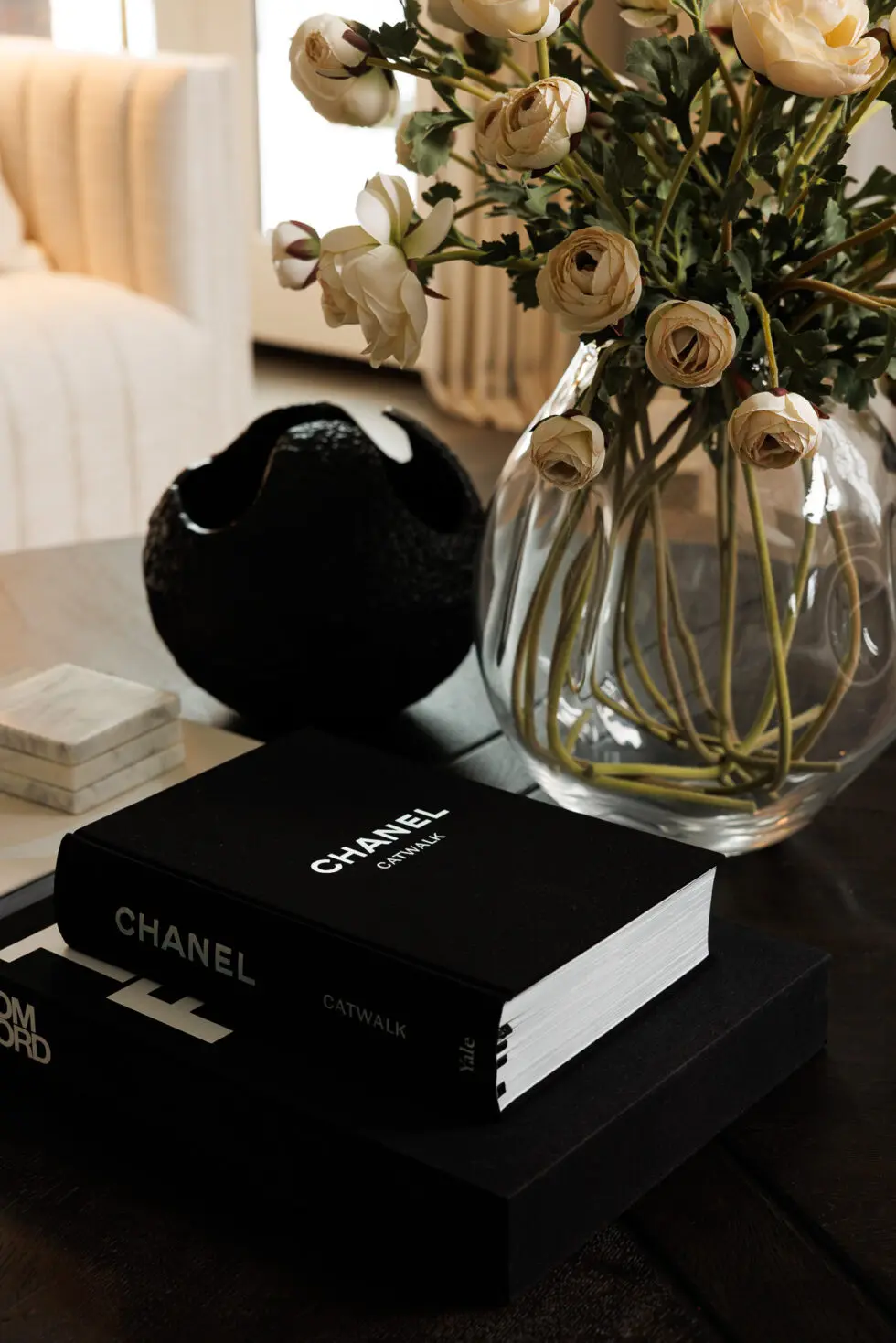 A black 'Chanel Catwalk' book stacked on another fashion book, likely 'Tom Ford', on a dark wooden coffee table, alongside a clear textured glass vase with light beige artificial ranunculus flowers, a black textured decorative bowl, and white marble coasters in a chic living room setting.