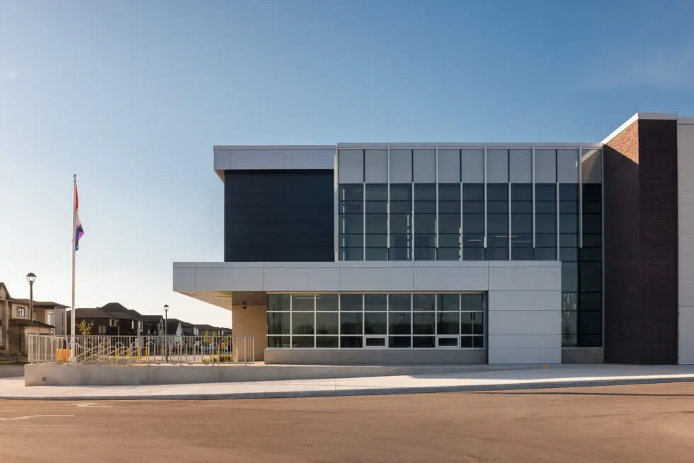 Modern public building with glass facade, grey panels, and brick accents under a clear blue sky, featuring a flag, bike racks, and an accessibility ramp.