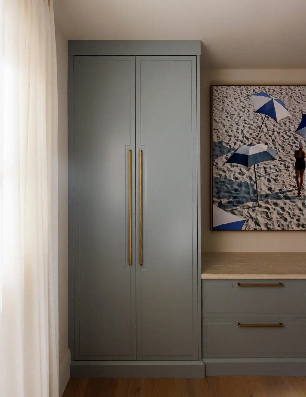 Elegant built-in grey wardrobe with brass handles, alongside a matching custom desk and drawers, and a large framed beach artwork with blue umbrellas.