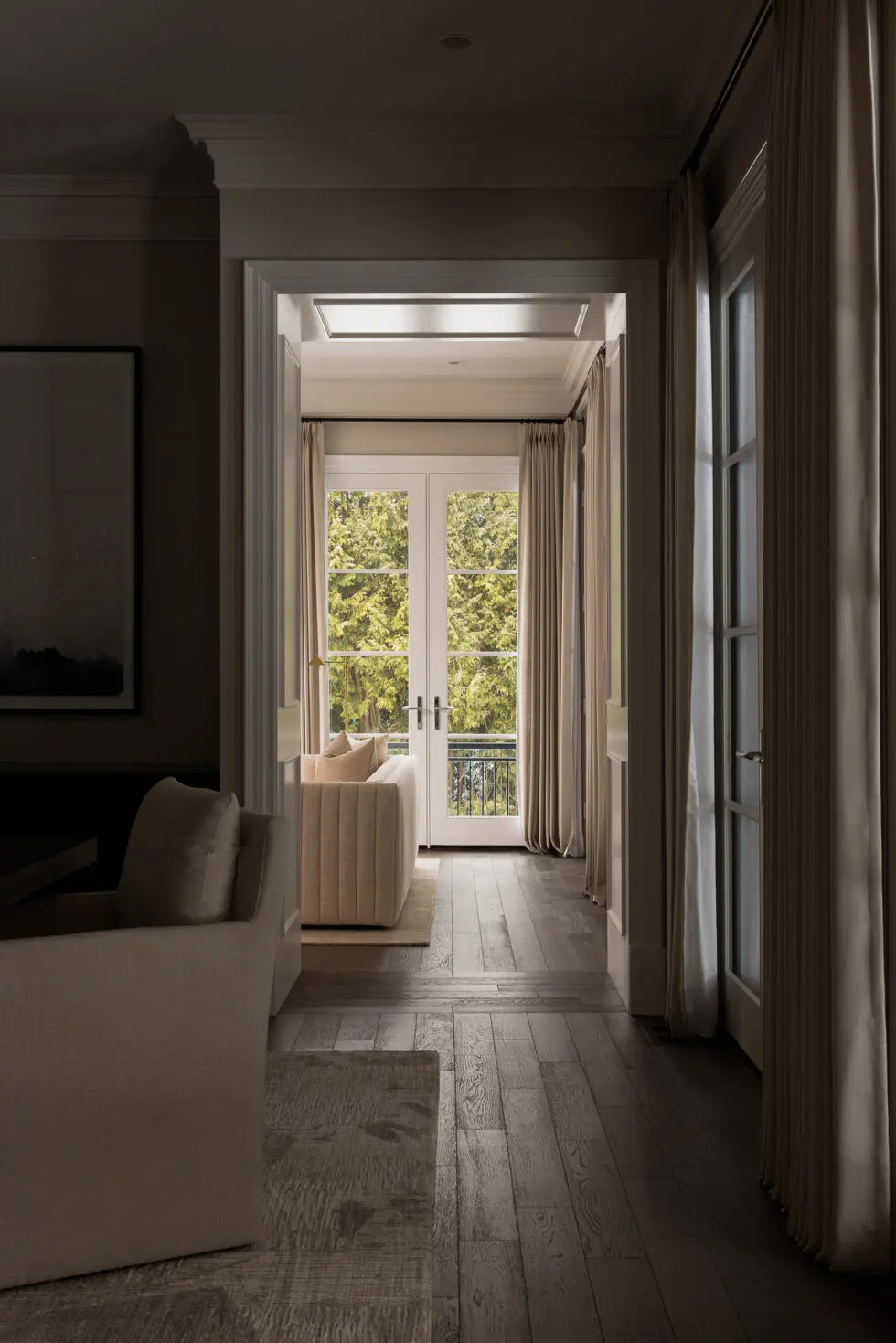 Elegant interior hallway with dark wood floors leading to a bright living room featuring French doors, a cream sofa, and beige curtains with a lush green outdoor view.