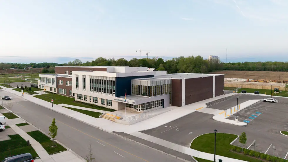 Aerial view of White Pine Public School at 3370, a modern building with white, dark grey, and brown facades, large glass windows, a spacious parking lot, and surrounding greenery under a clear sky.