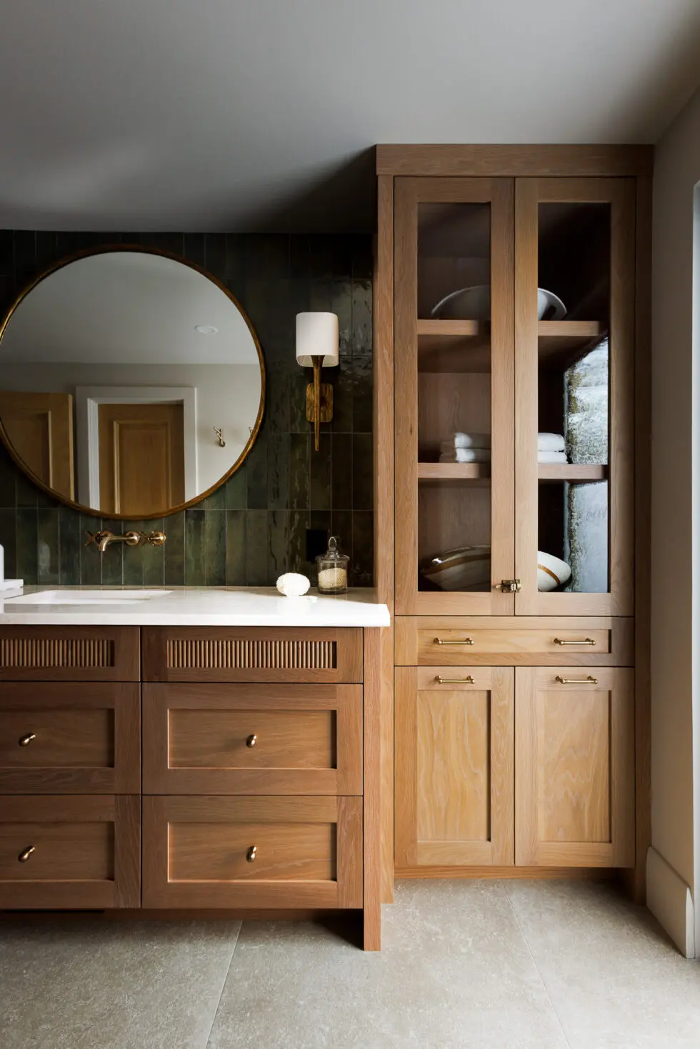 Modern bathroom with dark wood vanity, white counter, round brass mirror, green subway tiles, and light oak storage cabinet.