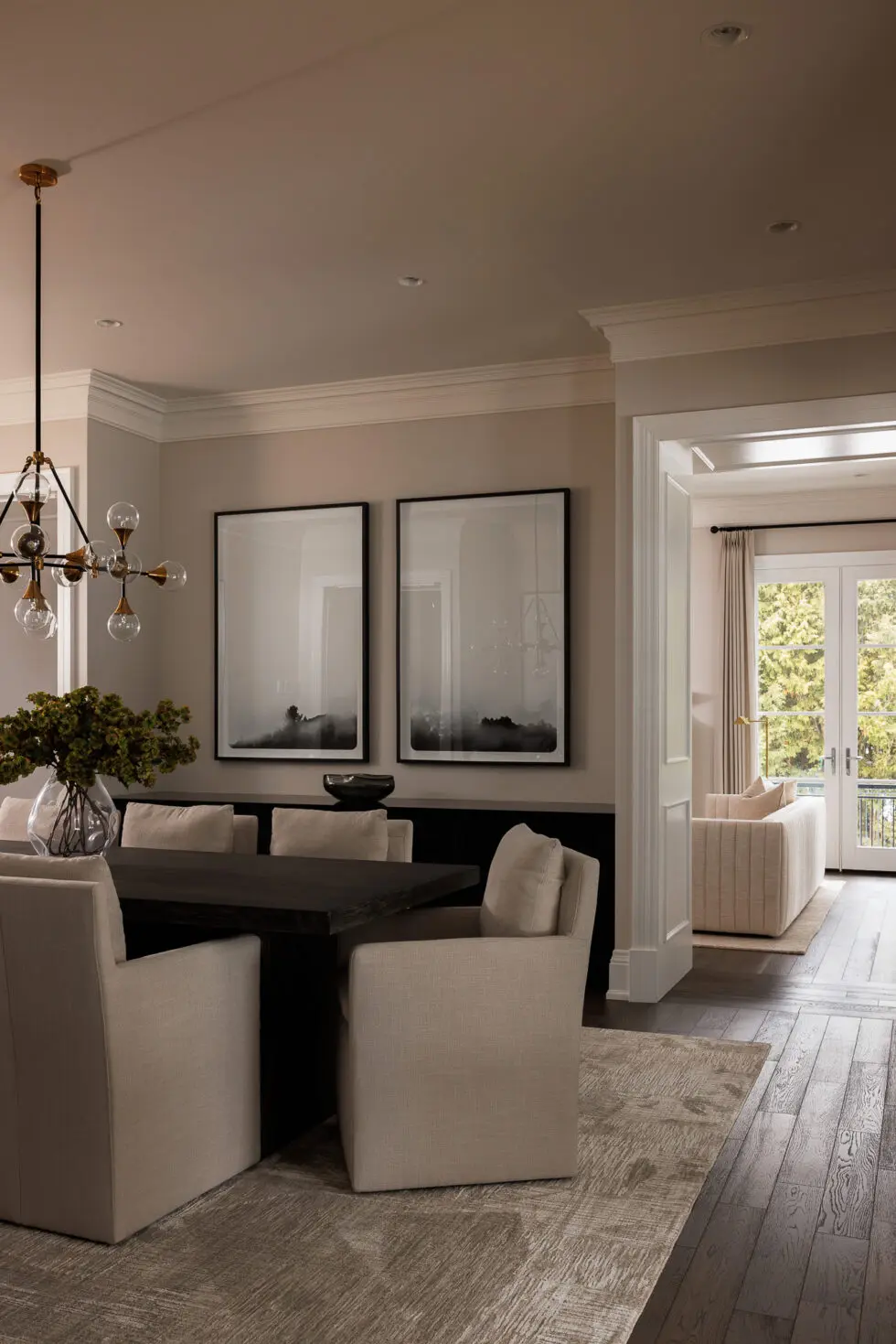 Modern dining room with a dark wood table, cream upholstered chairs, a brass and glass globe chandelier, and two large black and white abstract landscape artworks, leading to a living room with a cream sofa and French doors.
