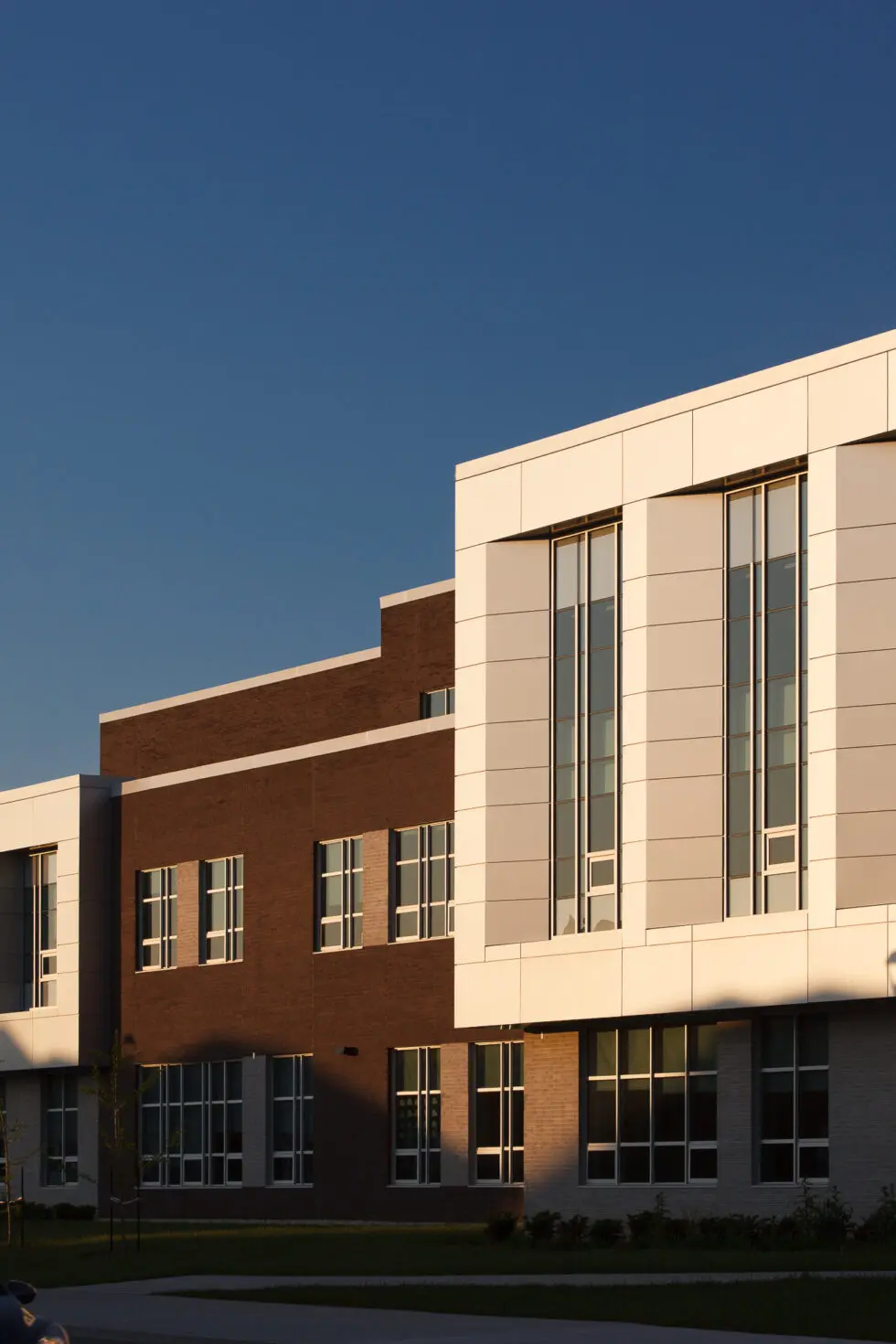 Modern building facade featuring white architectural panels and brown brick walls with numerous windows under a clear blue sky during golden hour.
