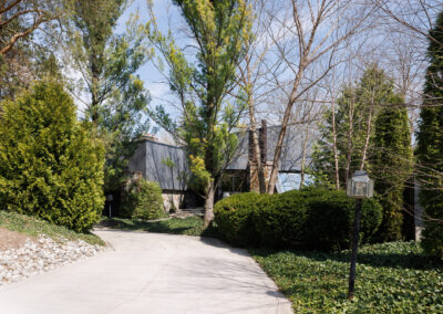 Modern house with dark grey facade and stone accents, surrounded by lush green trees and shrubs, viewed from a winding concrete driveway.