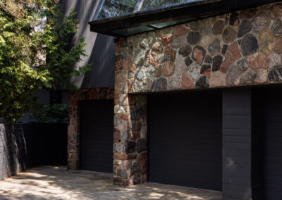 Modern architectural home featuring a natural stone facade, dark vertical wood siding, striking triangular glass windows, and multiple black garage doors, with a flagstone driveway in front.