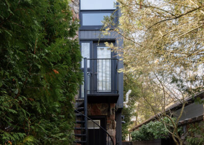 Modern narrow house exterior with stone facade, dark gray siding, and black spiral staircase leading to a balcony, surrounded by lush evergreen trees and a stone path.