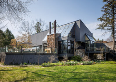Modern contemporary house with dark gray siding, extensive glass windows, stone accents, and a large outdoor deck set in a lush green yard.