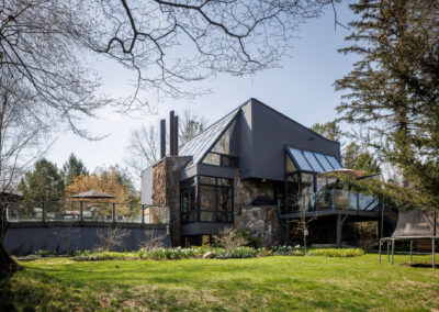 Modern angular house with dark siding, stone accents, extensive glass, and outdoor deck surrounded by a green lawn and trees in early spring.