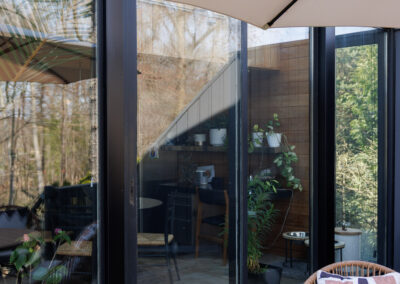 Modern outdoor patio with wicker chairs, round table, beige umbrella, and large glass doors reflecting trees and revealing an interior sunroom with plants.
