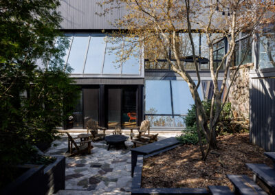Modern house exterior with an irregular stone patio, black fire pit, and wooden Adirondack chairs surrounded by trees.