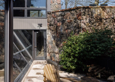 Outdoor patio with wooden Adirondack chairs on a flagstone surface next to a modern house with stone and glass architecture, surrounded by greenery.