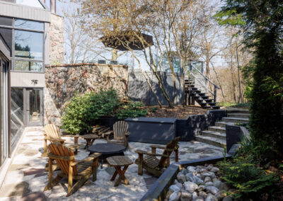 Outdoor flagstone patio with Adirondack chairs, fire pit, and multi-level landscaping next to a modern stone house.