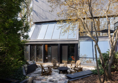 Modern house with a stone patio featuring a fire pit and Adirondack chairs, surrounded by lush trees and contemporary landscaping.