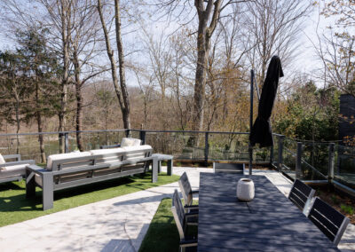 Modern outdoor patio deck with grey dining table, lounge seating, glass railing, and natural treed view overlooking water